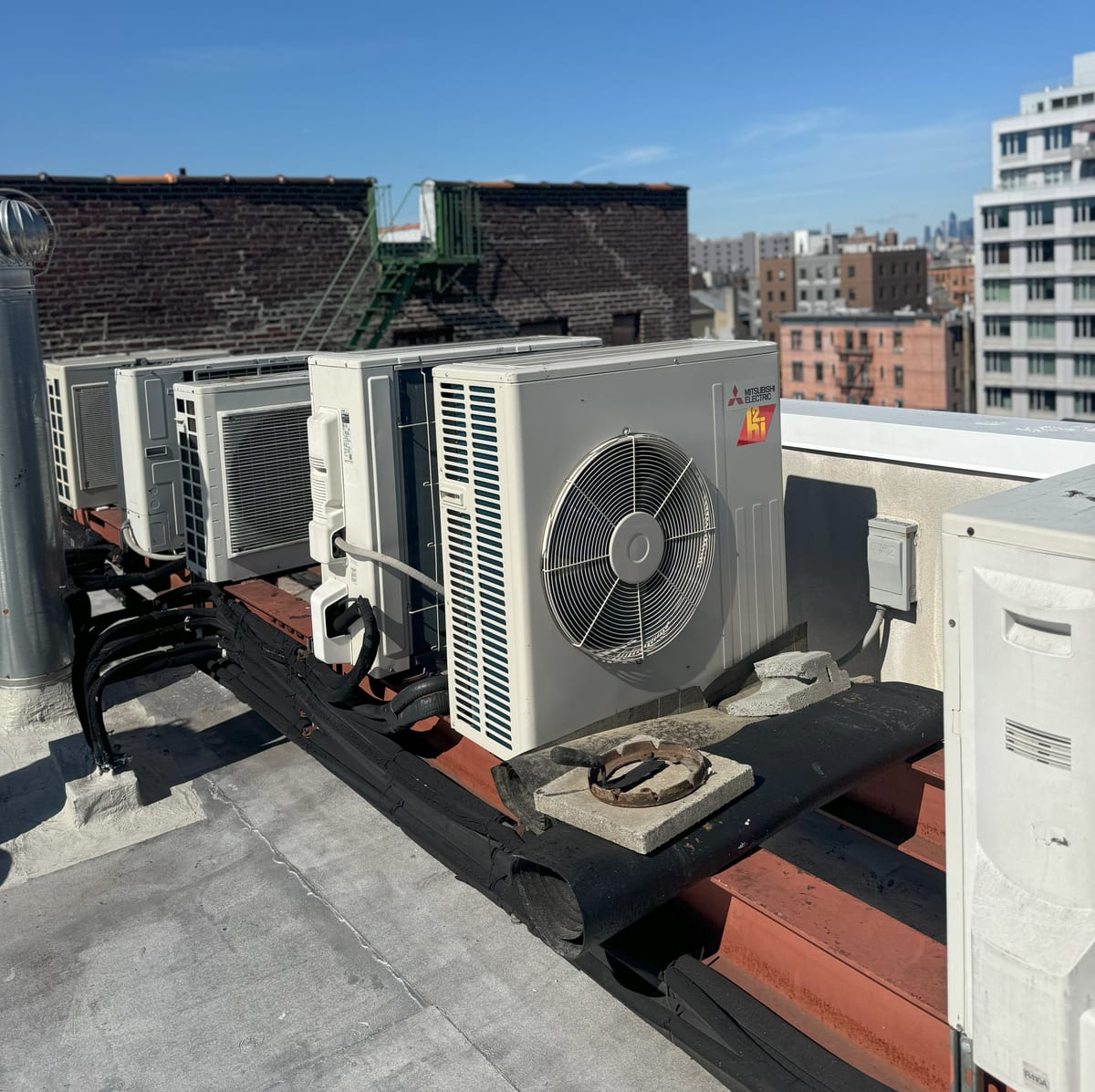 Row of Mitsubishi Electric Hyper-Heat (H2i) condensers on a Brooklyn rooftop with the New York City skyline behind